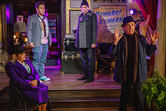 Four actors perform on a stage set featuring a man in a hat gesturing and a woman in a wheelchair.