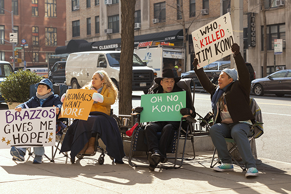 Four people sit on folding chairs on a city sidewalk holding various handmade protest signs.