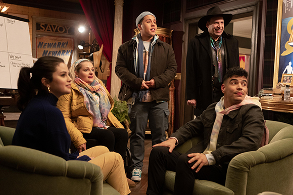 Five people in a room, some sitting in green armchairs and others standing, looking towards the left.