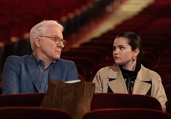 An older man in a blue blazer and a younger woman in a tan coat sit in red theater seats, looking at one another.