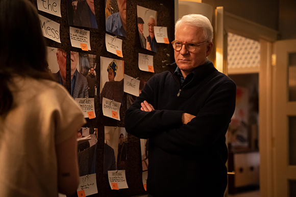 A man with white hair and glasses stands with arms crossed before a board filled with photographs and notes.