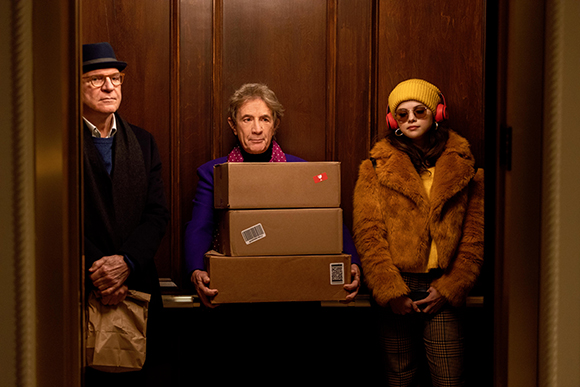 Three people stand in an elevator; one man holds stacked cardboard boxes, another holds a paper bag.