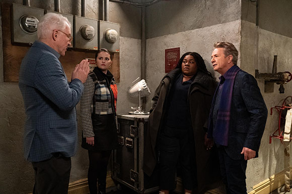 Four people stand in a dimly lit room with concrete walls, looking at one another with serious expressions.