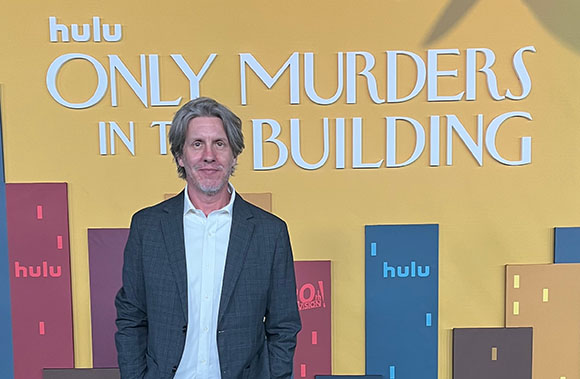 A man in a grey blazer and white shirt stands before a yellow Hulu promotional backdrop.