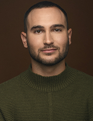Headshot of a man with short dark hair and a beard wearing an olive green ribbed sweater against a brown background.