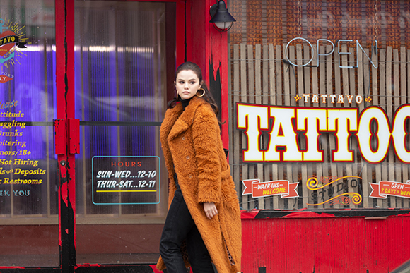 A woman in a long brown fuzzy coat stands before a red storefront with a large tattoo studio sign.