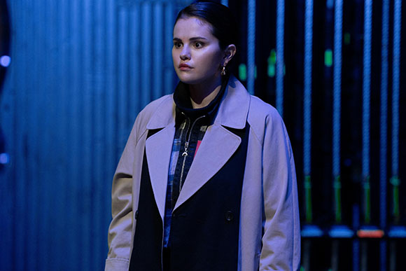 A young woman with dark hair wearing a light beige coat looks pensively to the side against a blue background.
