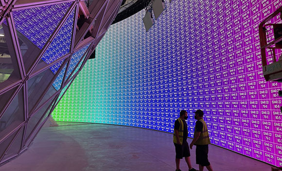 Two workers in high-visibility vests stand before a large, curved LED wall with a blue and purple grid pattern.