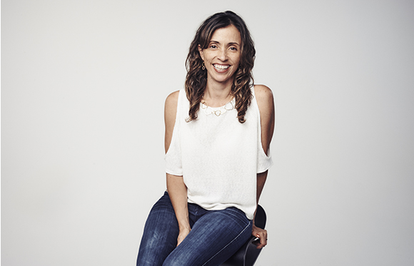 A smiling woman with long wavy hair wearing a white sleeveless top and blue jeans, sitting against a white background.