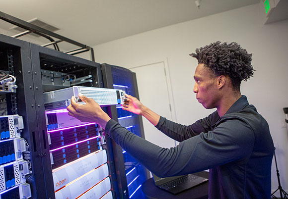 A man in a black long-sleeve shirt installs a silver server component into a glowing blue data center rack.