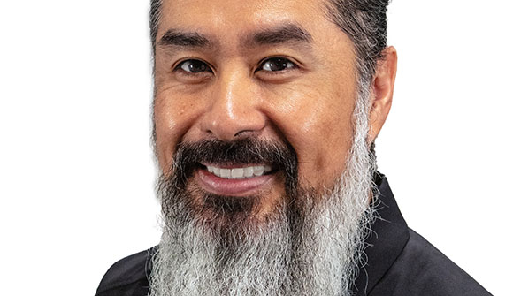 A smiling man with dark hair and a long, thick grey and black beard against a white background.