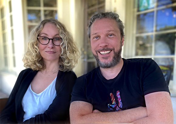 A woman with blonde curly hair and glasses and a man with a beard smiling side by side.