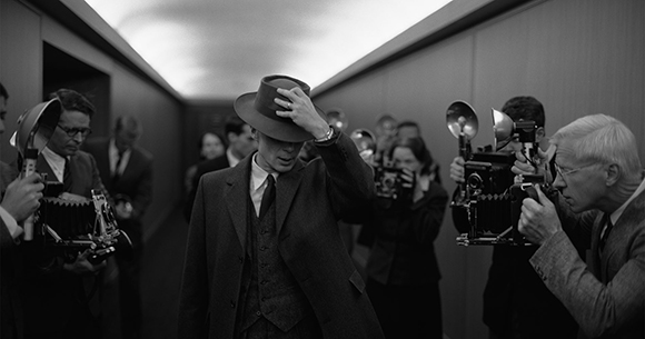 A man in a suit and fedora walks through a crowd of people holding cameras.