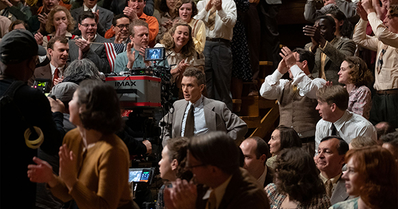 A film crew with an IMAX camera films a large crowd of people in period clothing clapping in a theater.