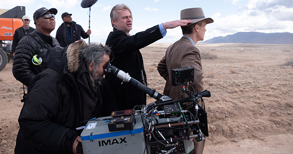 A film crew works with a large IMAX camera in a vast, dry desert landscape under a cloudy sky.