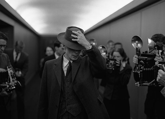 A man in a suit and fedora adjusts his hat while photographers with vintage cameras surround him.