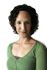A woman with dark curly hair and a green v-neck shirt smiling slightly against a white background.