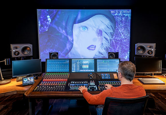 A person sits at a large mixing desk in a dark studio facing a massive screen displaying anime art.