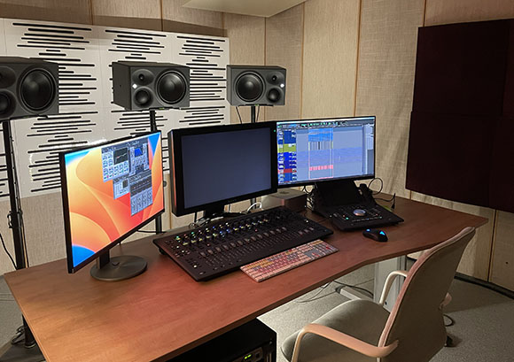 A music studio desk with two computer monitors, a mixing console, a keyboard, and studio monitors on stands.