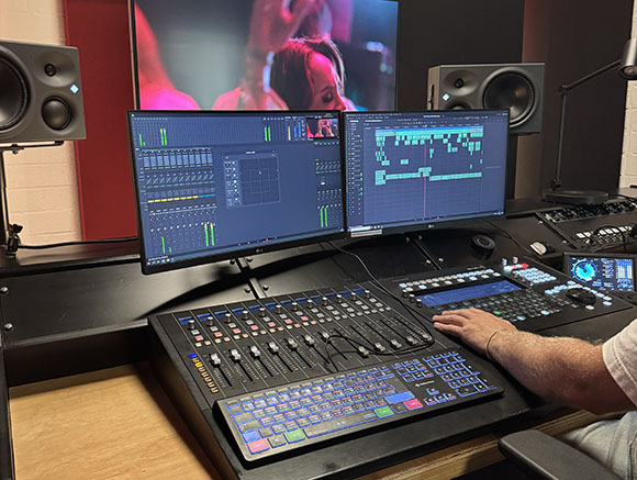 A person's hand operates a large digital audio mixing console in a studio with dual monitors and speakers.