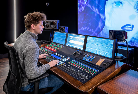 A man sits at a professional audio mixing desk with multiple monitors in a dark studio.