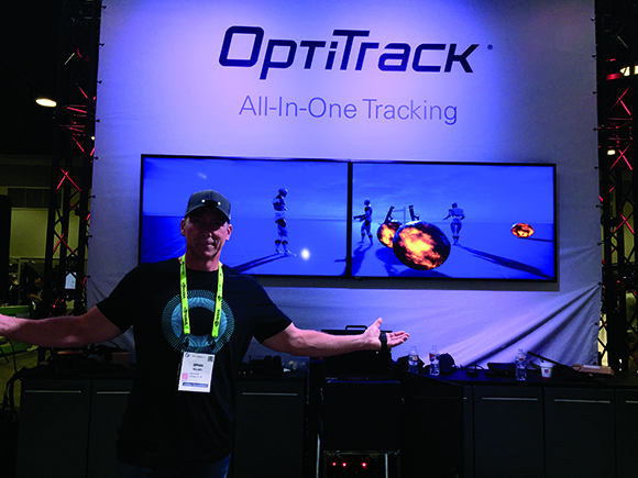 A man gestures toward a large OptiTrack screen displaying motion capture graphics at an exhibition.