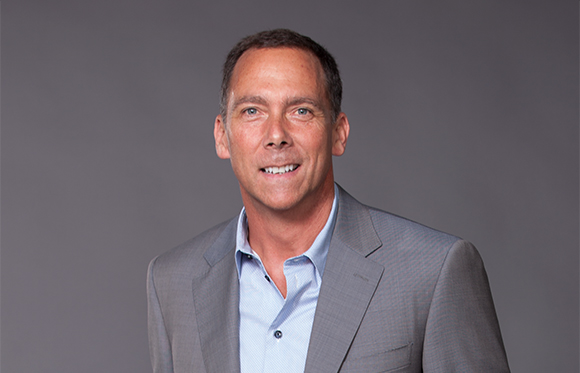 A smiling man with short dark hair wearing a light blue shirt and a grey suit jacket.