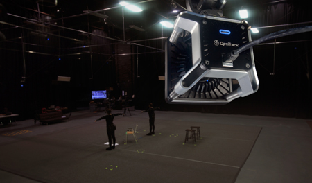 A high-tech OptiTrack motion capture camera in the foreground overlooking a dark studio with people in motion suits.