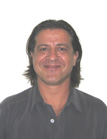 A smiling man with dark, shoulder-length hair wearing a dark grey collared shirt against a white background.