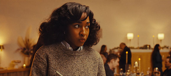 A woman with dark wavy hair and a grey sweater looks to the side in a warm, dimly lit room.