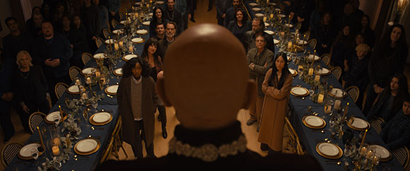 A person's bald head in the foreground overlooks a long, crowded dining table in a dimly lit room.