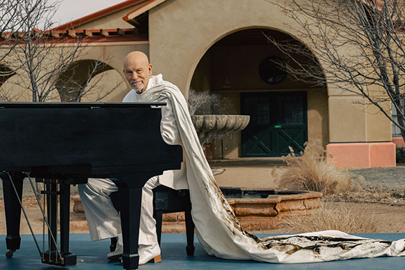 A bald man in white robes sits behind a black grand piano in front of a tan building.