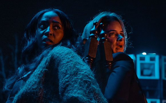 Two women with expressions of fear looking off-camera in dark, blue-toned lighting.