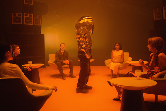 A person in a reflective metallic suit stands in a room among seated people under orange light.