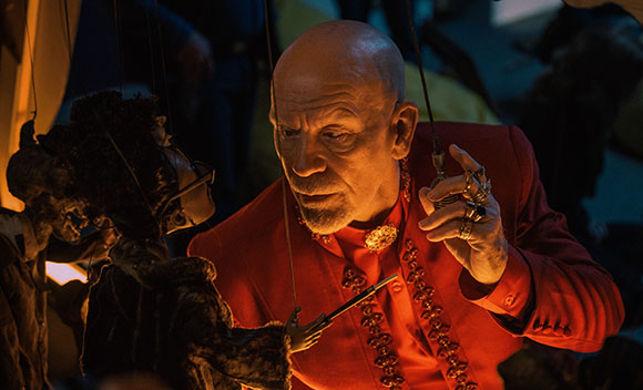 A bald man in a red ornate jacket manipulates a dark puppet with thin strings.