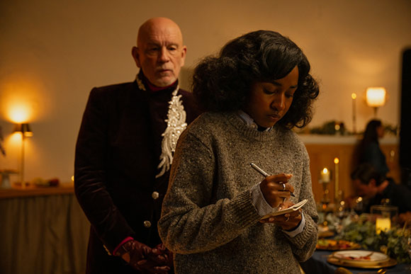 A woman in a grey sweater writes in a notebook while a man in a dark velvet jacket stands behind her.