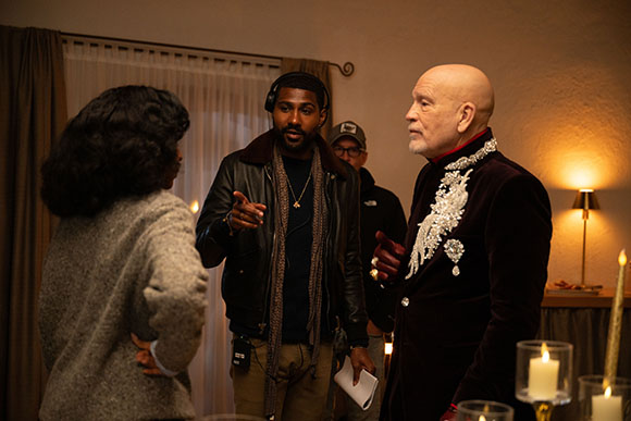 A man in a dark leather jacket gestures toward a woman while talking to a bald man in a velvet jacket.