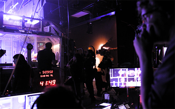 Film crew silhouettes working in a dark studio illuminated by purple light and glowing monitors.