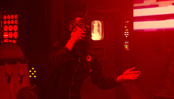 A person wearing goggles and a dark jacket gestures with one hand in a room bathed in red light.