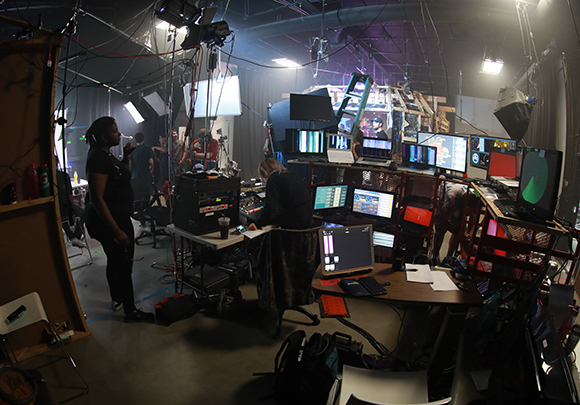Film crew in a dark studio surrounded by monitors, lighting rigs, and production equipment.