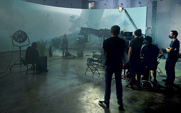 Film crew members standing in a studio before a large curved screen displaying a misty forest landscape.