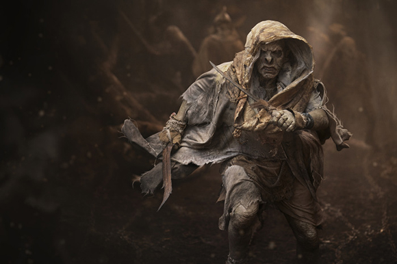 A pale, hooded figure in ragged, earth-toned clothing holds a small blade in a dark, misty environment.