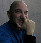 A bald man in a blue shirt smiles while resting his chin on his hand against a gray wall.