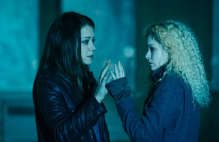 Two young women stand facing each other in a dimly lit, blue-toned environment.