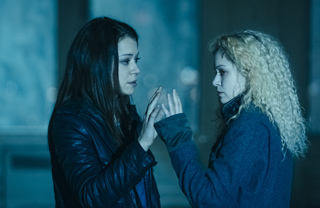 Two young women stand facing each other with their hands raised between them in a dimly lit setting.
