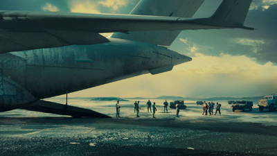 Rear view of a large cargo plane on a runway with military personnel and vehicles in the background.