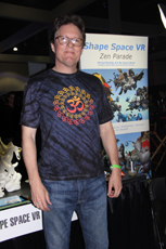 A man wearing glasses and a tie-dye t-shirt with an Om symbol stands in front of a booth promoting Shape Space VR Zen Parade.