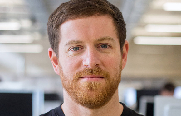 Close-up of a man with reddish-brown hair and beard, blue eyes, and a slight smile, looking directly at the camera. The backg
