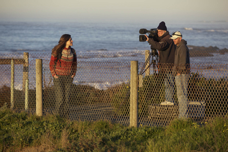 A woman in a red sweater stands by a fence as a man films her with a professional camera near the ocean.