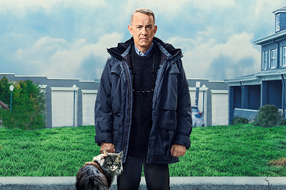 A man in a dark navy parka stands behind a fluffy grey cat on a grassy lawn.